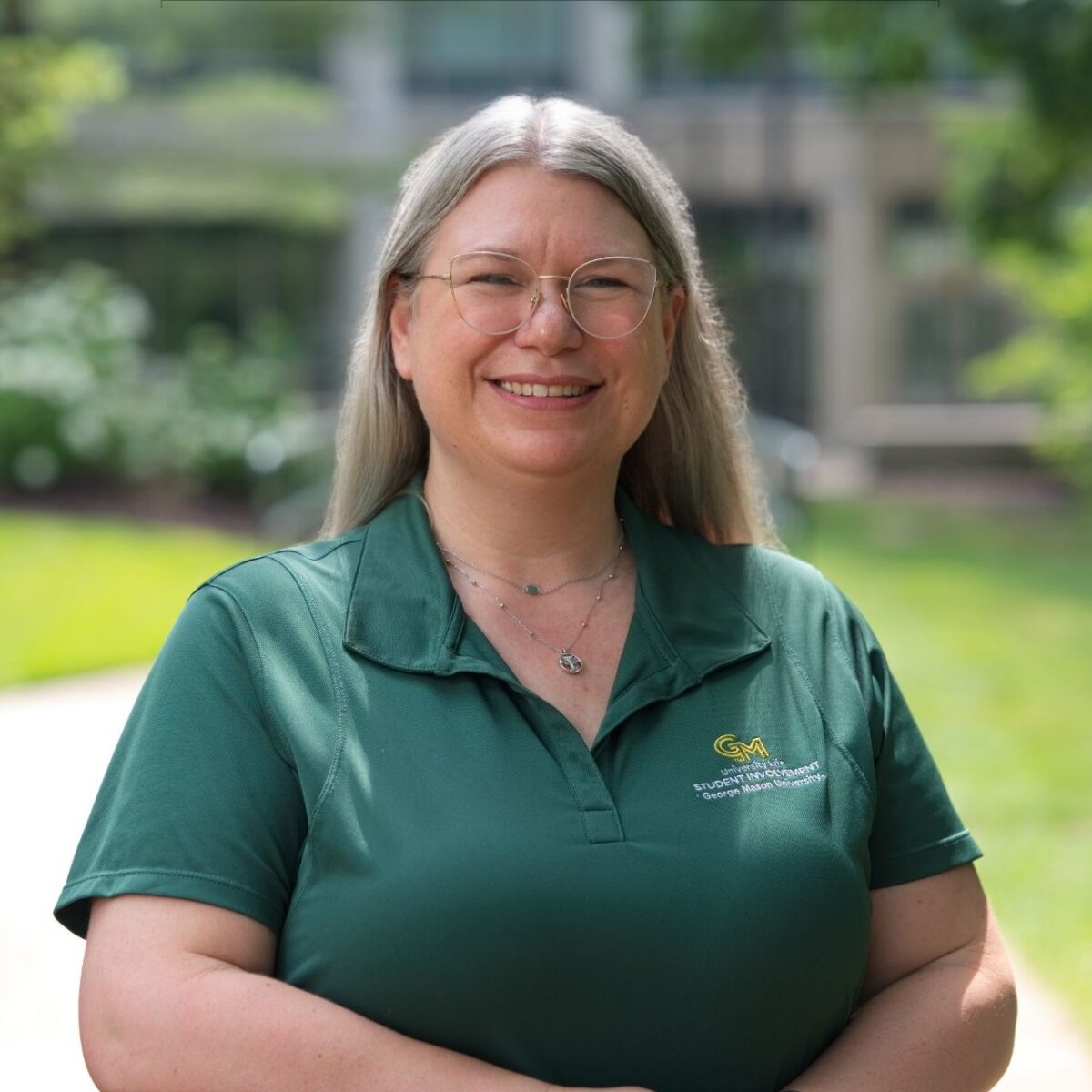 Professional outdoor headshot of a smiling person with long gray/blonde hair and glasses wearing a dark green George Mason University Student Involvement polo shirt. The person faces the camera with a blurred campus setting in the background.