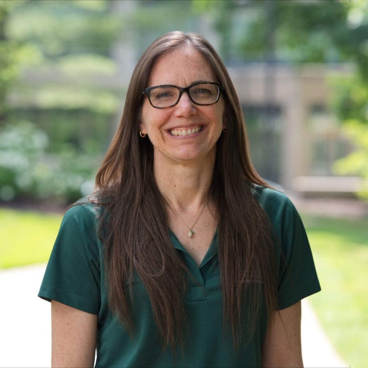 A professional portrait of a smiling person standing outdoors on a college campus. They have long brown hair, wear black rectangular eyeglasses, small hoop earrings, and a dark green polo shirt. The background is softly blurred, showing a walkway, trees, and campus buildings in natural daylight.