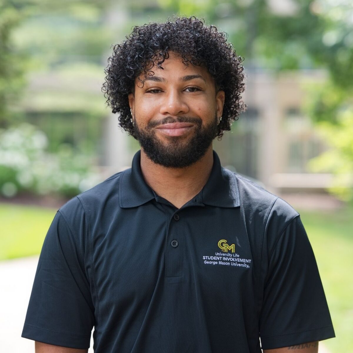 Professional outdoor headshot of staff member "Kenyatta", with short curly brown hair wearing a dark green George Mason University Student Involvement polo shirt. The person faces the camera with a blurred campus walkway and greenery in the background.