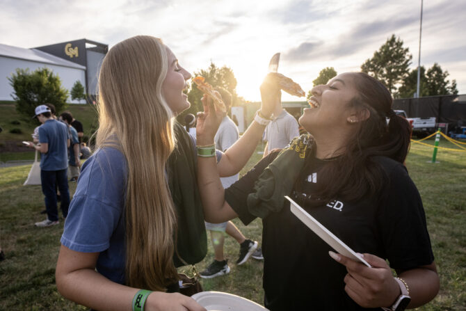 Two students laugh while eating slices of pizza outdoors at a George Mason University event. One student playfully holds a slice toward the other as they stand together on a grassy field, with other attendees and campus facilities in the background. Warm sunlight shines behind them.